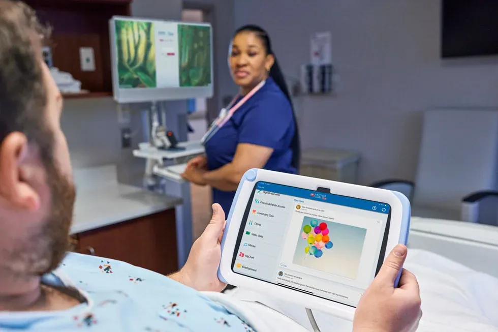 A hospital patient using the MyChart Bedside app on an iPad while a nurse works at a bedside computer cart