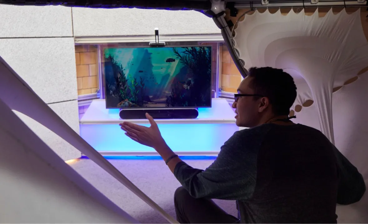 A student sitting in a mesh cocoon gesturing towards interactive screen displaying an underwater scene with purple mood lighting