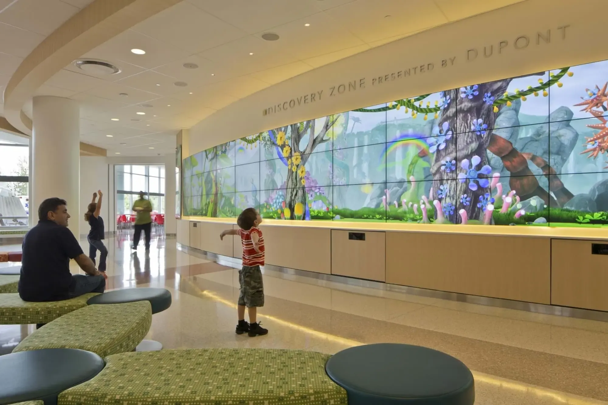A child standing with arms wide open in front of a large interactive video wall in a pediatric hospital