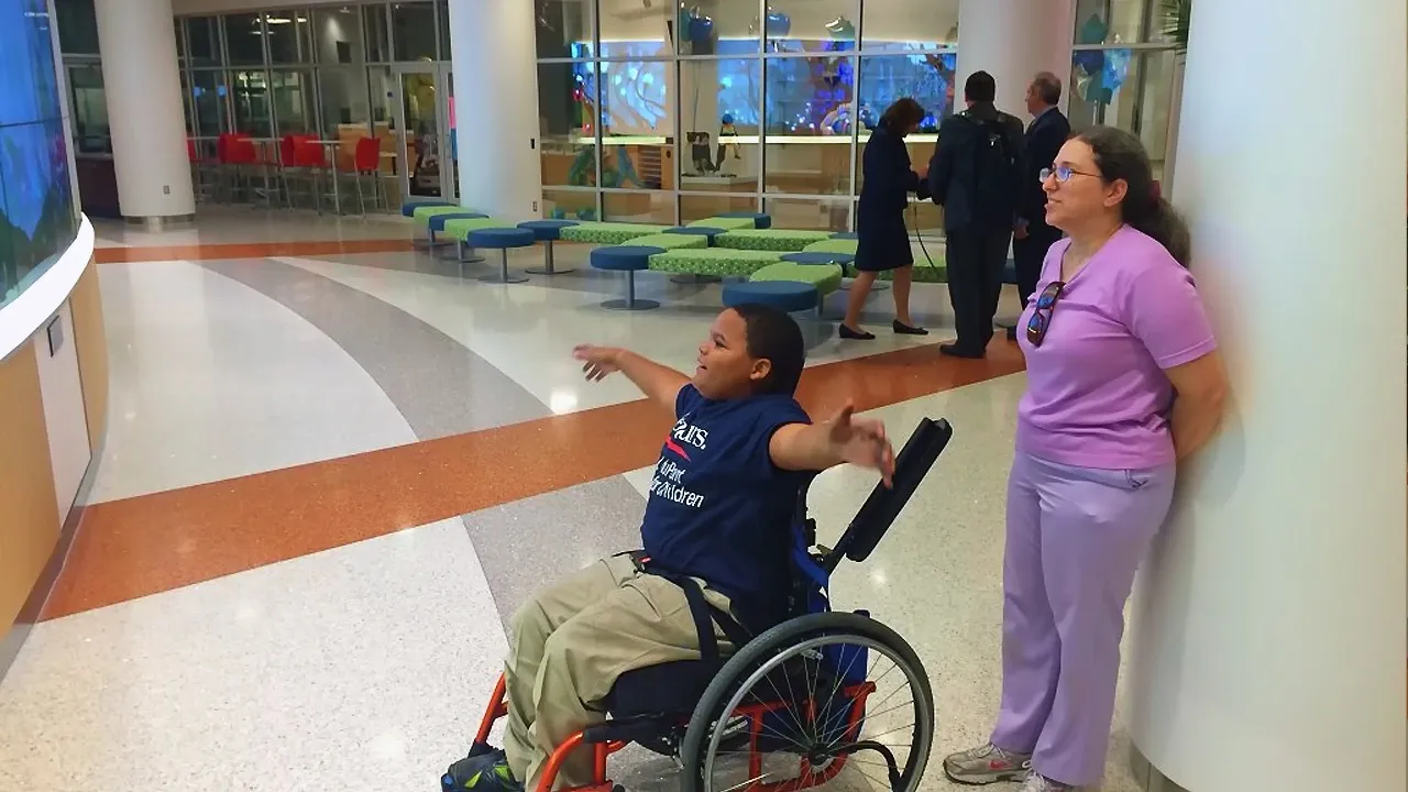 A kid on wheelchair gesturing towards a large interactive video wall, smiling with arms wide open. Behind, a nurse is leaning against a column watching him.